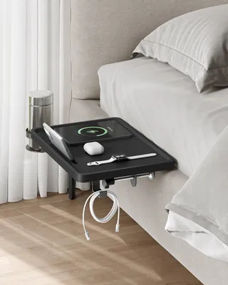 A black bedside table attached to a bed, holding various electronic devices and a drink.