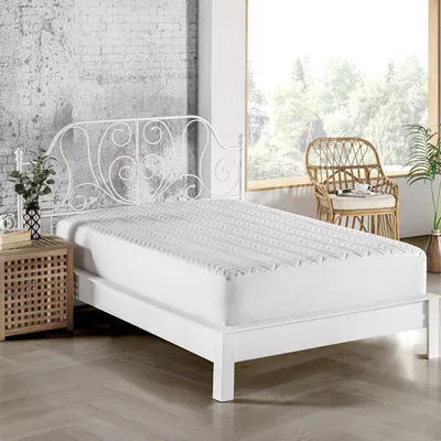 A white bed with a quilted mattress topper is shown in a bedroom setting with a window and chair in the background.