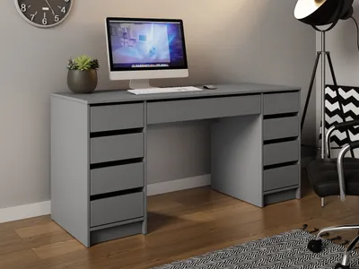 A gray desk with a computer and a plant on it, set in a room with a clock on the wall and a floor lamp and chair in the background.