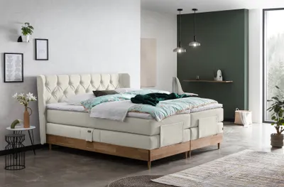 A bedroom scene featuring a large, adjustable boxspring bed with a tufted headboard, dressed in bedding. The room has minimalist decor with a dark green accent wall and a window.
