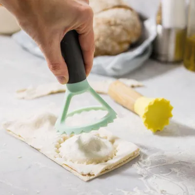 A hand is using a green ravioli cutter to seal a ravioli on a floured surface. Another ravioli cutter with a yellow head and wooden handle is visible in the background.