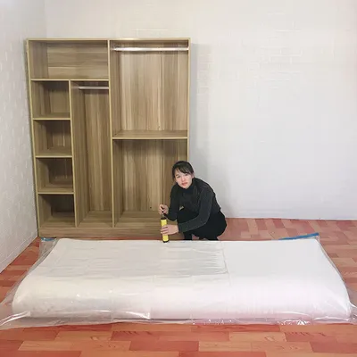 A woman is crouching next to a large, vacuum-sealed mattress bag in front of a wooden wardrobe.