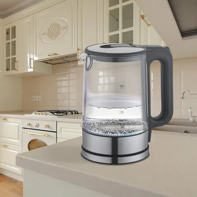 A glass electric kettle filled with water is shown on a kitchen countertop, with kitchen cabinets and a stove in the background.