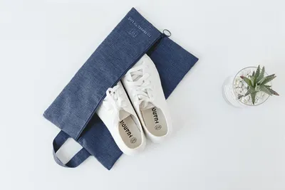 A pair of white sneakers is placed inside a blue denim organizer bag, which is laid open on a white surface. A small potted succulent plant is visible to the right.
