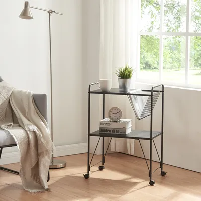 A small black rolling cart with two shelves is placed next to a gray armchair with a blanket draped over it. A floor lamp stands behind the armchair. A window with white curtains is visible in the background.