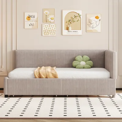 A daybed with a mattress, a yellow blanket, and a green flower-shaped pillow is shown in a room with artwork on the wall and a patterned rug on the floor.