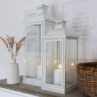 Two white lanterns with lit candles are displayed on a wooden surface next to a vase with dried grass and a woven basket.