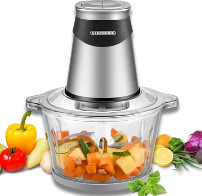A food chopper with chopped vegetables inside, surrounded by fresh vegetables and herbs.