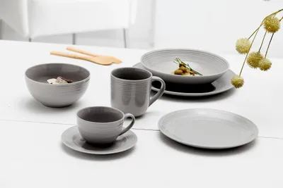A set of grey ceramic tableware, including bowls, mugs, and plates, arranged on a white wooden table.