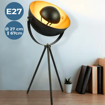 A black tripod floor lamp with a gold interior shade is shown against a light gray and white wall, with books and a small plant on a wooden surface in the foreground.
