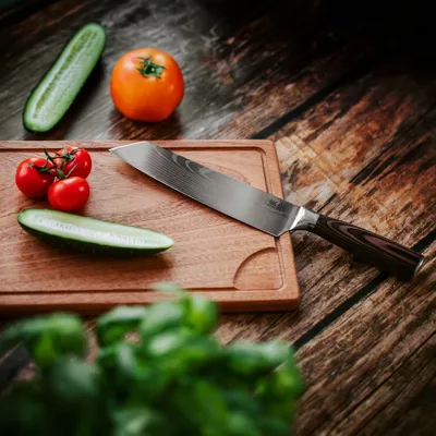 A kitchen knife is placed on a wooden cutting board with vegetables like tomatoes and cucumbers.