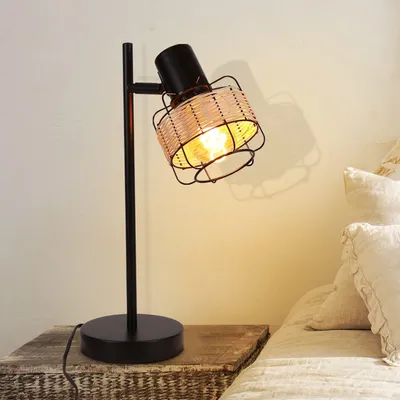 A table lamp with a rattan shade sits on a bedside table next to a bed.