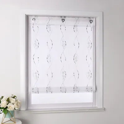 A white roller blind with a gray embroidered pattern is shown hanging in a window frame. A small bouquet of white flowers in a teal vase is on a table to the left.