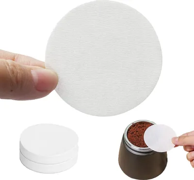 A hand holding a round coffee filter paper, with a stack of filters and a coffee maker with coffee grounds in the background.