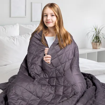 A young girl is sitting on a bed, wrapped in a gray weighted blanket.