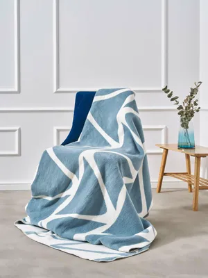 A blue and white geometric patterned blanket draped over a chair in a room with white paneled walls and a small wooden side table with a vase of greenery.