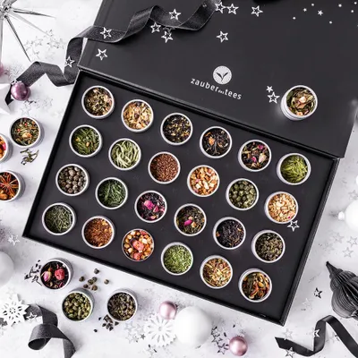 A black gift box filled with small tins of tea, decorated with stars and ribbon, on a white and gray speckled surface with festive elements like snowflakes and ornaments.