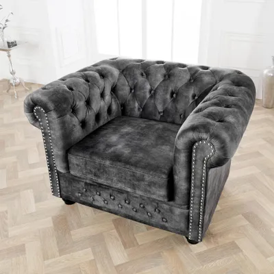 A dark gray velvet armchair with button tufting and silver nailhead trim, placed on a light wood floor in a well-lit room.