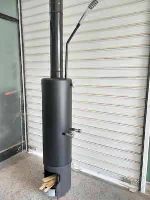A black cylindrical wood-burning boiler with a chimney and a small opening at the bottom containing firewood, set against a white roll-up door.