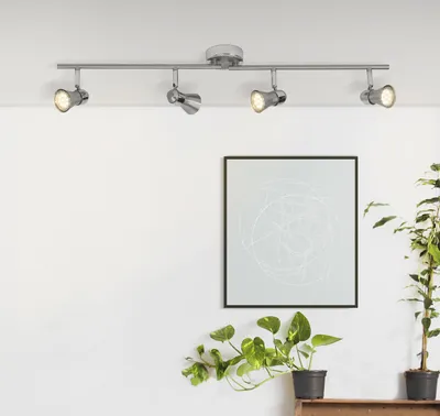 A track lighting fixture with four adjustable spotlights mounted on a ceiling, with a framed abstract artwork and potted plants on the wall below.