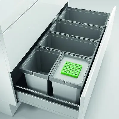 A pull-out drawer from a kitchen cabinet containing multiple waste bins, one of which has a green insert with multiple small compartments.