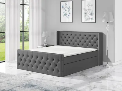 A dark gray upholstered boxspring bed with a tufted headboard and footboard, set up in a bedroom with large windows and artwork on the wall.