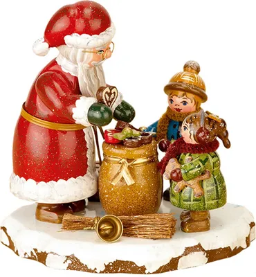 Zu sehen ist eine weihnachtliche Figurenszene mit einem Weihnachtsmann der zwei Kindern Geschenke überreicht während sie auf einem schneebedeckten Sockel stehen und ein Besen sowie eine Glocke den Boden schmücken