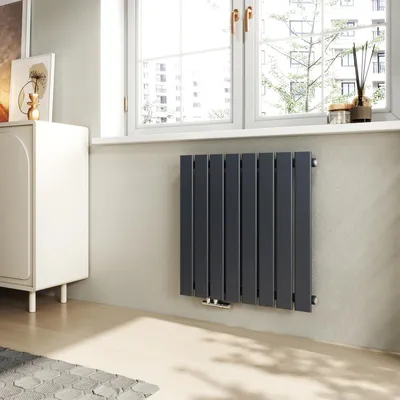 A modern anthracite radiator is mounted on a light gray wall next to a white cabinet, with a window in the background.