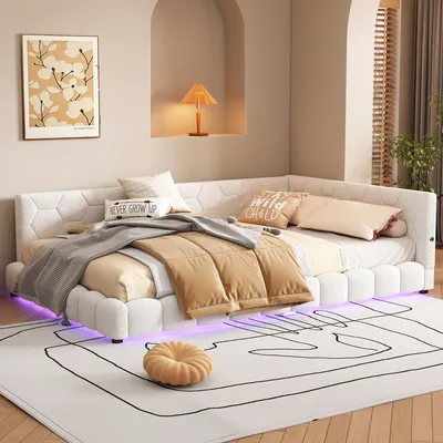 A modern white upholstered corner bed with decorative pillows and throws, set in a bedroom with soft lighting and a wall niche containing a lamp.