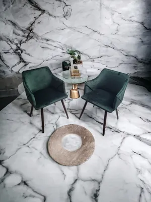 Two green velvet chairs are placed on a marble floor in front of a marble wall, with a small round table between them.