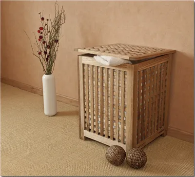 A wooden laundry hamper with a lid, a white vase with dried flowers, and two woven balls on a carpeted floor, next to a wall.