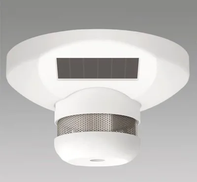A white smoke detector with a solar panel on top and a metallic grille on the bottom, shown against a light gray background.
