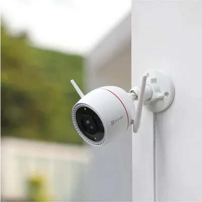 A white security camera with two antennas is mounted on a white wall. The background is blurred green foliage.