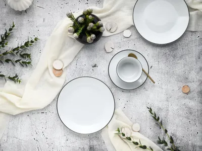 A flat lay of a white porcelain coffee service set with black trim, arranged on a textured gray surface with a cream-colored fabric runner and some greenery.