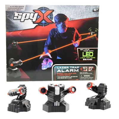 The image shows the packaging of a SpyX Lazer Trap Alarm toy, with a boy demonstrating its use. Below the packaging are three individual units of the toy.