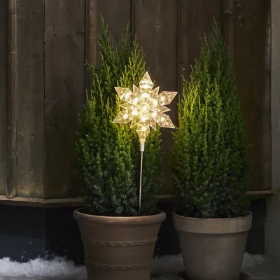 A snowflake-shaped light decoration is placed between two potted evergreen trees in front of a wooden wall.
