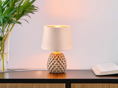 A table lamp with a textured ceramic base and a fabric shade sits on a dark wooden surface next to a vase of plants and a book.