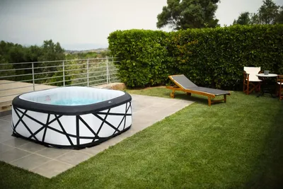An inflatable hot tub is set up on a patio next to a grassy lawn, with a lounge chair and table in the background.