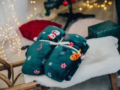 Christmas-themed blankets tied together, placed on a fluffy white surface, with a Christmas tree and lights in the background.