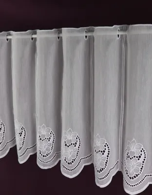 A close-up view of white lace curtains with floral embroidery along the bottom edge, hanging from a curtain rod against a dark purple background.