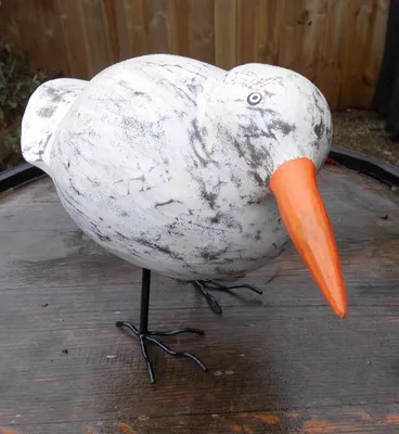 A decorative wooden bird sculpture with an orange beak and black wire legs, placed on a wooden surface.