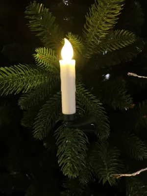 A close-up shot of a white LED candle with a flame-like light, clipped onto a green Christmas tree branch.
