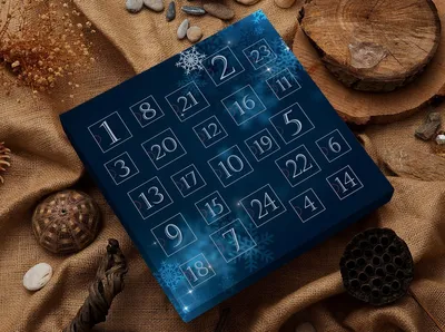 An advent calendar with numbered squares is placed on a burlap fabric background, surrounded by natural elements like stones, wood, and dried plants.