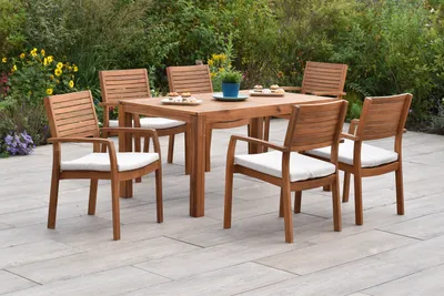 A wooden garden dining set with six chairs and a table is set up on a patio, surrounded by greenery and flowers.