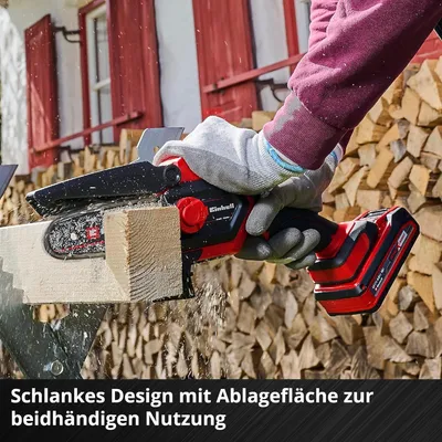 A person is using a cordless chainsaw to cut a piece of wood, with a stack of firewood in the background.