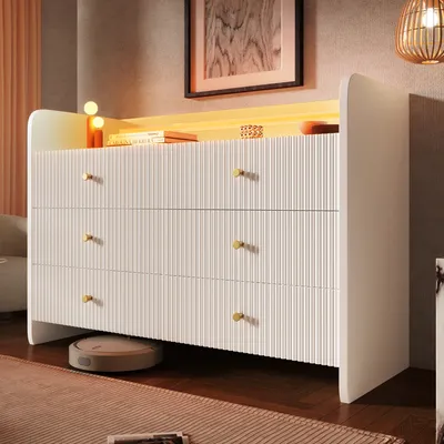 A white dresser with gold knobs is shown in a room with warm lighting. A robot vacuum cleaner is visible underneath the dresser.
