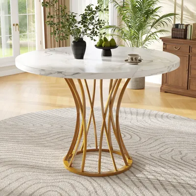 A round marble-top dining table with a gold pedestal base is set in a well-lit dining room with plants and a wooden cabinet in the background.