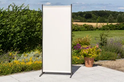 A beige privacy screen stands on a paved patio in a garden setting.