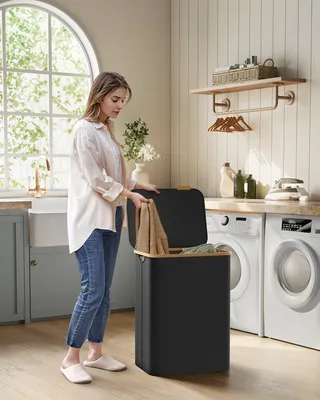A woman is putting laundry into a black hamper with a wooden lid in a laundry room.
