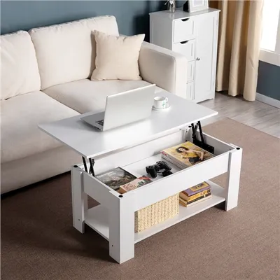 A white lift-top coffee table is shown in a living room setting, next to a sofa. The table has a laptop, a cup, books, and a game controller on it.